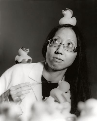 Anita is pictured in a lab coat in a black and white photo, trying to observe a crowd of rubber ducks to no avail. One of them sits on her head.