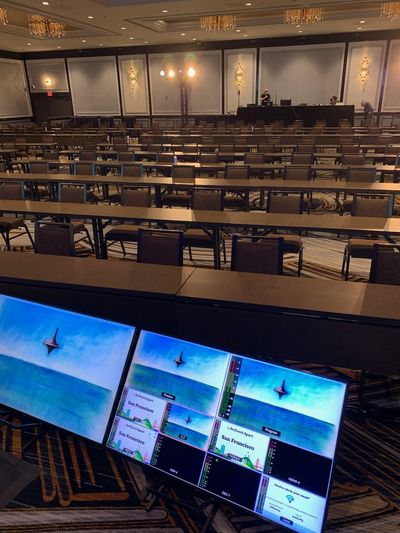 The view from the stage at tech check, looking out at rows and rows (and rows) of empty chairs at tables, with multiple screens of my laptop background displaying on the two monitors as well as “An Event Apart” branded screens.