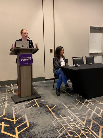 Two speakers at the front of the room: an Asian woman in a blazer and jeans sitting at a table looking offscreen at slides, and a blonde white woman in a blazer standing at the lectern with a laptop while speaking.