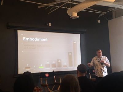 A man presents a slide with the content, 'Embodiment: What kinds of bodies do screen sci-fi AI have?' with a bar chart where 'humanoid' is almost as many as all the other options (which includes virtual, vehicular, and mechanical) combined.'