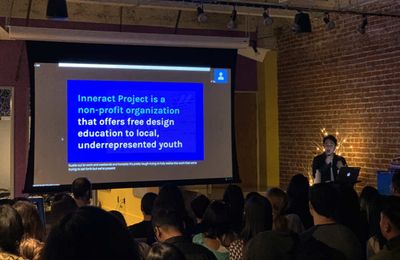 Jihern speaks into a mic in front of a slide with the content, 'Inneract Project is a non-profit organization that offers free design education to local, underrepresented youth.'