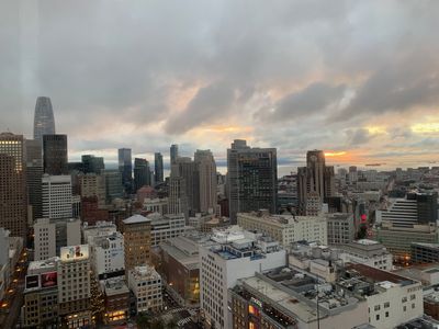 From a hotel highrise, the sun peeks through clouds above the San Francisco Bay, with downtown San Francisco buildings seen in the foreground.