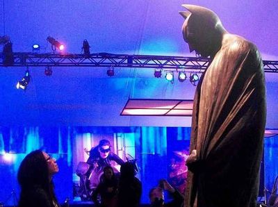 An Asian woman stands in a dramatically lit presentation hall and looks up at a very tall statue of Batman, wrapped in his cape, from the film The Dark Knight Rises.