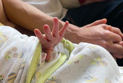 A newborn's tiny hand stretches out from inside a swaddle, as her parents hold hands in the background.