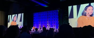 Four panelists sit on a brightly lit stage on low couches, with two large screens on either side showing one panelist, an Asian-Latina woman wearing yellow.