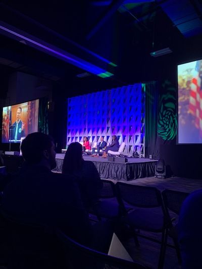 Three panelists sit on a brightly lit stage on low couches, with two large screens on either side showing another speaker at a lectern in a fancy briefing room.