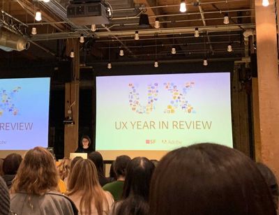 A photo taken from the middle of an industrial-looking common space, looking toward the stage where there are two large screens with "UX Year in Review" displayed.